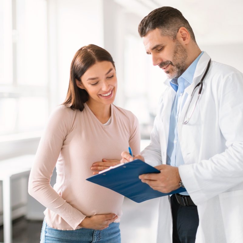 Male doctor showing clipboard with medical test results to happy pregnant lady during meeting in